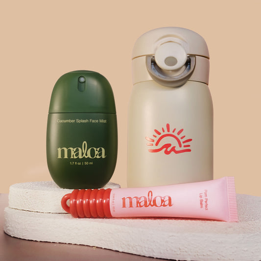 Three MALOA skincare/cosmetic products arranged on a textured white surface against a warm beige background: a dark green face mist bottle - Cucumber Splash Face Mist, a cream insulated tumbler with a coral sun logo, and a pink tube with a red cap - Pom Perfect Lip Balm