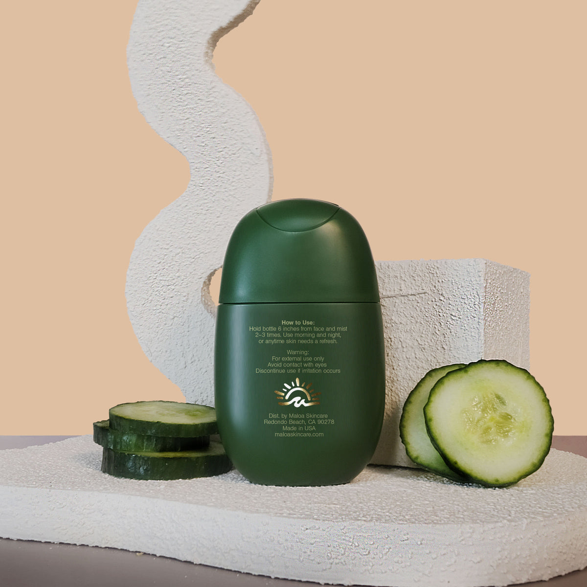 The back of a matte dark green bottle of maloa Cucumber Splash Face Mist sits on a textured white stone, flanked by fresh cucumber slices, with a beige background and abstract decorative props. Back exposing gold foil maloa logo