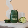 A matte dark green bottle of maloa Cucumber Splash Face Mist sits on a textured white stone, flanked by fresh cucumber slices, with a beige background and abstract decorative props.