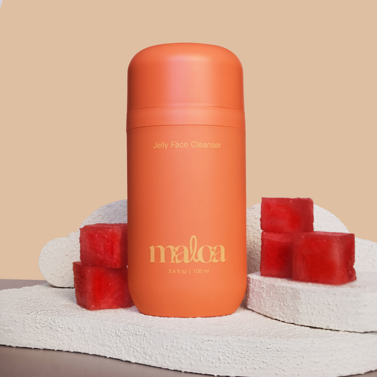 An terracotta bottle of MALOA Jelly Face Cleanser stands centered on a textured white stone slab, flanked by watermelon cubes against a warm beige background.