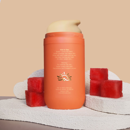 The back of a terracotta bottle of MALOA Jelly Face Cleanser stands centered on a textured white stone slab exposing the back with gold foil Maloa logo, flanked by watermelon cubes against a warm beige background.