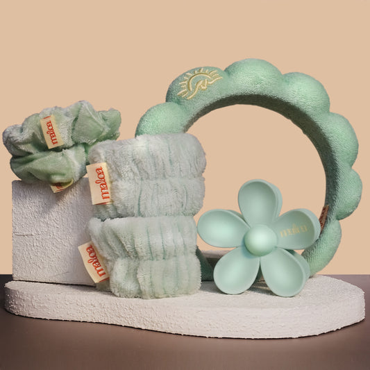 A MALOA branded self-care set displayed on a pedestal against a warm beige background. The set includes pale mint green wristies, two matching scrunchies, a flower-shaped hair clip, a spa headband