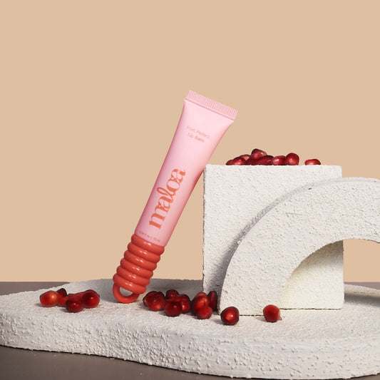 A pink tube of MALOA Pom Perfect Lip Balm standing on a textured stone platform, surrounded by scattered and stacked pomegranate seeds