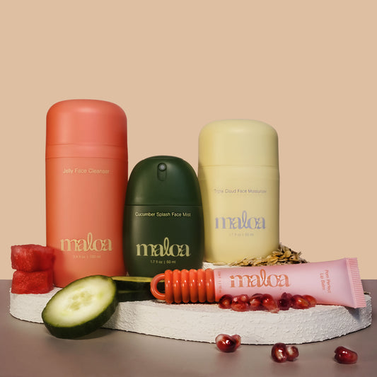 Four MALOA skincare products displayed on a white stone slab with natural ingredients: a coral pink Jelly Face Cleanser bottle, a dark green Cucumber Splash Face Mask bottle, a pale yellow Triple Cloud Face Moisturizer bottle, and a pink tube of Pom Perfect Lip Balm, arranged with watermelon, cucumber, oats and pomegranate on a beige background.