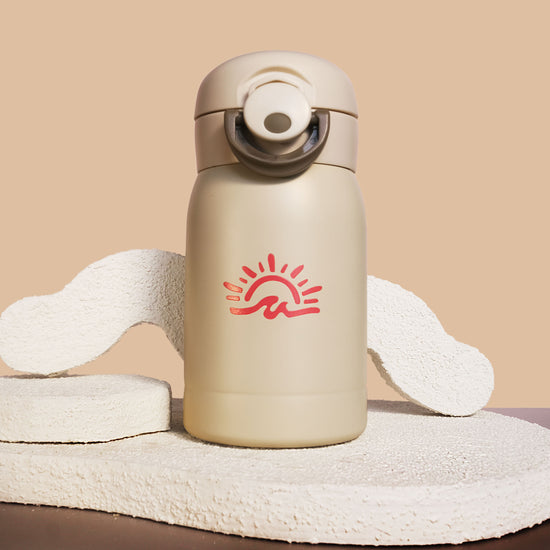 A cream-colored insulated water bottle with a red sun and wave logo, displayed on a textured beige stone prop against a solid beige background.