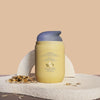 The back of a pale yellow Triple Cloud Face Moisturizer with the brand name 'MALOA' printed in blue on the front, styled on a textured stone surface with scattered oats against a warm beige background. Back exposing the metalic gold foil logo