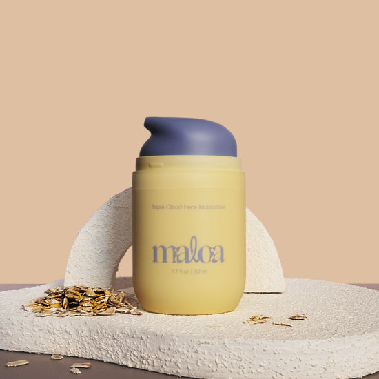 A pale yellow Triple Cloud Face Moisturizer with the brand name 'MALOA' printed in blue on the front, styled on a textured stone surface with scattered oats against a warm beige background. Top off exposing blue pump.