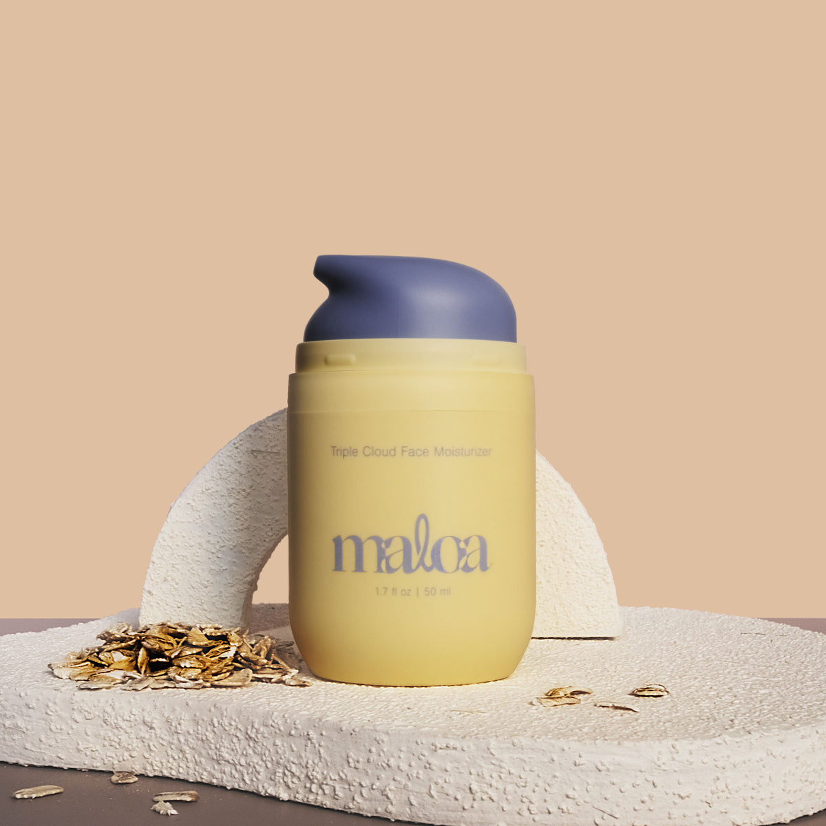 A pale yellow Triple Cloud Face Moisturizer with the brand name 'MALOA' printed in blue on the front, styled on a textured stone surface with scattered oats against a warm beige background. Top off exposing blue pump.