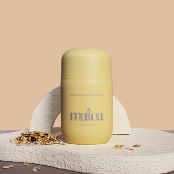 A pale yellow Triple Cloud Face Moisturizer with the brand name 'MALOA' printed in blue on the front, styled on a textured stone surface with scattered oats against a warm beige background.