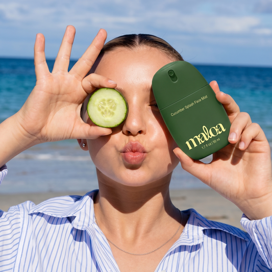 A woman on a beach holds a dark green Maloa Cucumber Splash Face Mist bottle in one hand and a cucumber slice in the other, with the ocean and sky in the background.