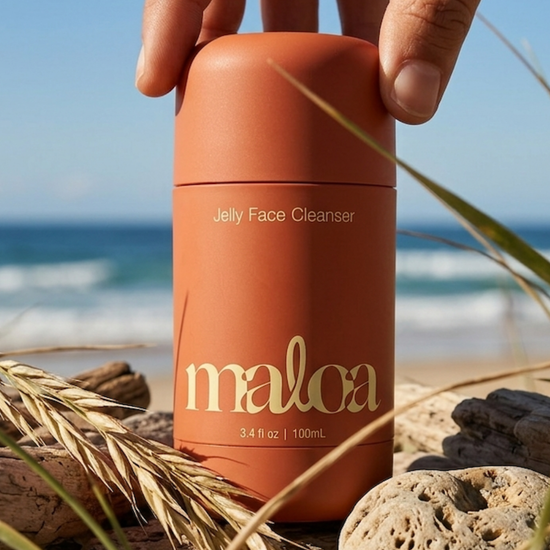 A hand holds a coral orange cylindrical bottle of Maloa Jelly Face Cleanser on a beach with rocks, sand, and ocean waves in the background.
