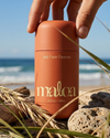 A hand holds a coral orange cylindrical bottle of Maloa Jelly Face Cleanser on a beach with rocks, sand, and ocean waves in the background.
