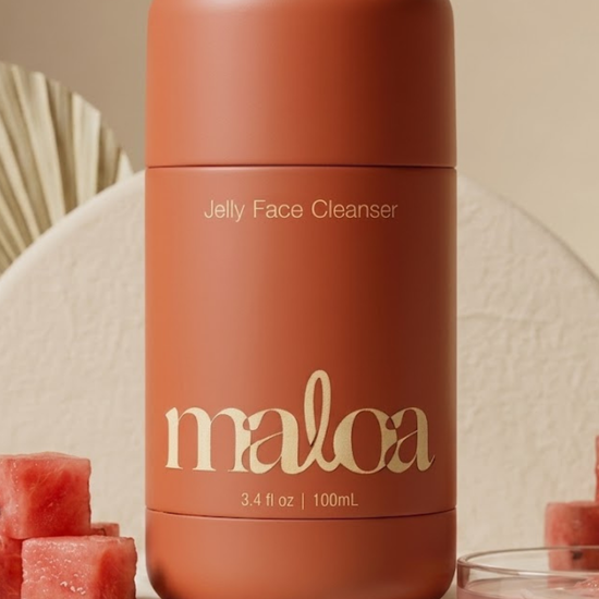 A matte terracotta-colored bottle of maloa Jelly Face Cleanser, styled on a textured beige surface with watermelon cubes and seeds.