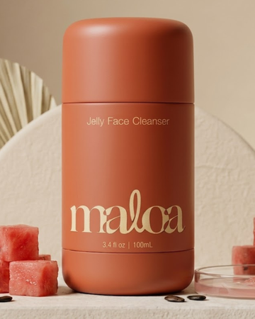 A matte terracotta-colored bottle of maloa Jelly Face Cleanser, styled on a textured beige surface with watermelon cubes and seeds.