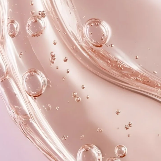 A close-up of a glossy, translucent pink cosmetic gel or liquid with floating air bubbles on a soft pink background.