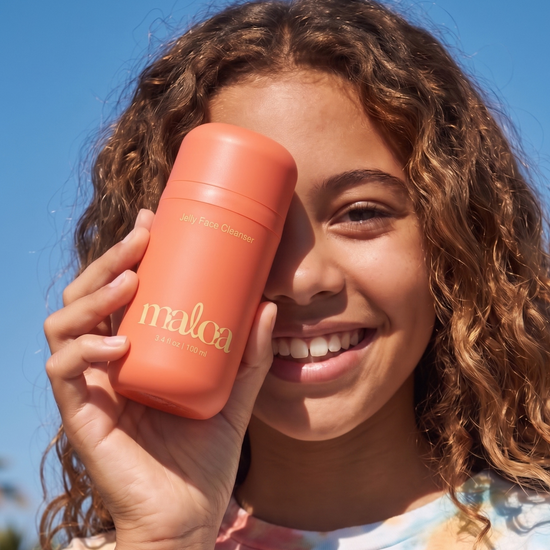 A smiling tween outdoors holds a coral-pink cylindrical bottle labeled 'maloa jelly face cleanser' in front of her face against a bright blue sky.