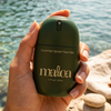 A hand holding a dark green bottle of Maloa Cucumber Splash Face Mist against a blurred background of water and rocks.