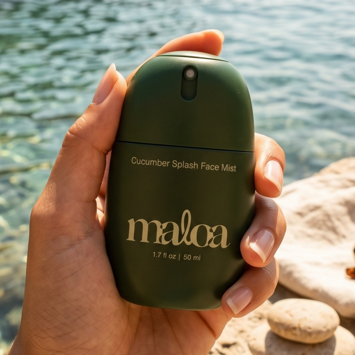 A hand holding a dark green bottle of Maloa Cucumber Splash Face Mist against a blurred background of water and rocks.
