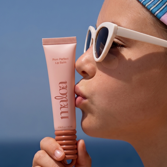 A close-up lifestyle shot of a tween wearing white cat-eye sunglasses and a blue striped headband, holding a pink squeeze tube of 'maloa Pom Perfect Lip Balm' to her lips against a clear blue sky and ocean background.