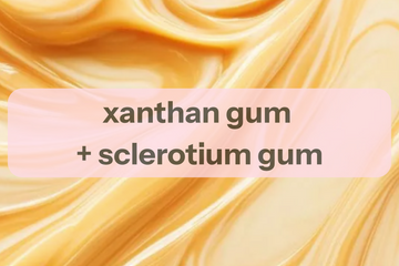 A digital graphic with a background of golden-yellow marbled liquid texture, featuring a semi-transparent pink rounded rectangle overlay with the text 'xanthan gum + sclerotium gum' in dark gray.